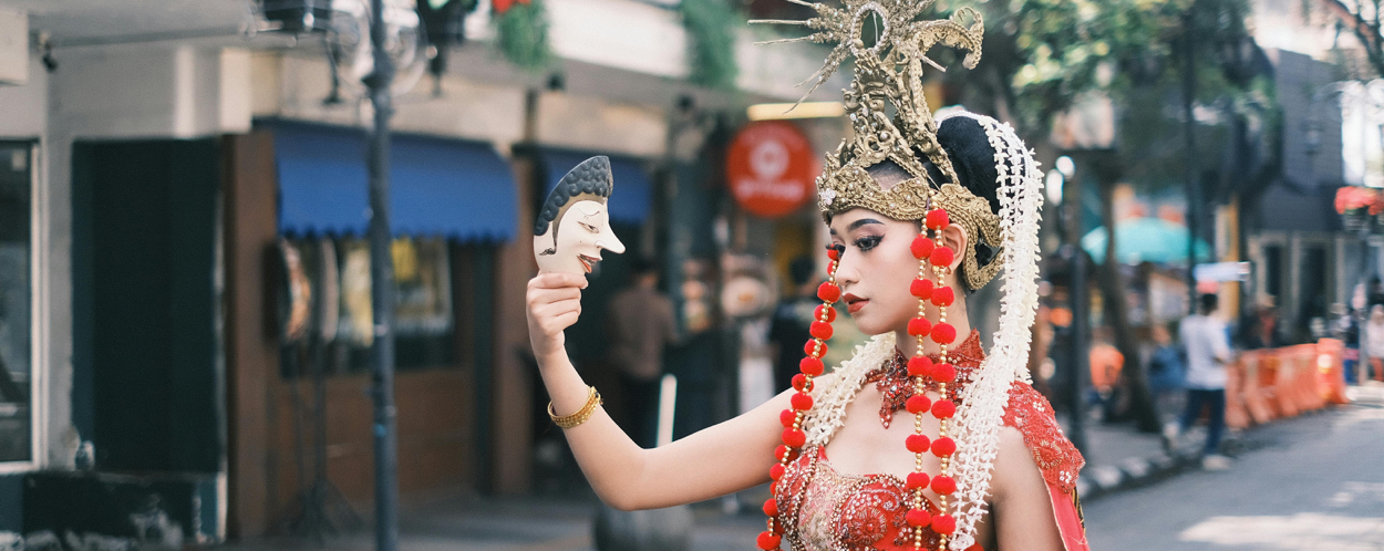 Eine traditionelle Tänzerin in reich verziertem Kostüm und Kopfschmuck hält eine Maske in einer Straße in West-Java, Indonesien.